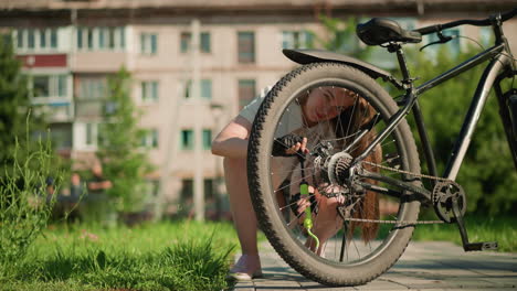 young woman with warm smile crouches on paved path, inflating rear tire of bicycle with air pump, surrounded by greenery and blurred background of building and people walking by