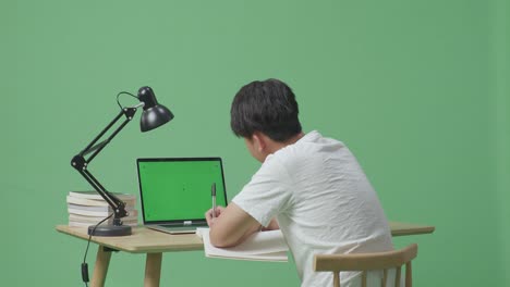 젊은 아시아 남성이 녹색 스크린 노트북을 보고 집에서 녹색 화면 배경에서 공부하는 동안 책에 글을 쓰고 있습니다.