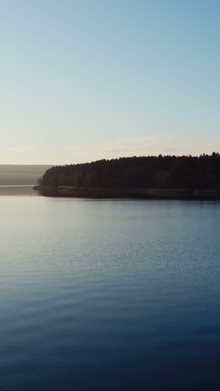Flying over blue water in the evening. Beautiful calm river on trees background in nature at sunset. Motion camera forward. Vertical video
