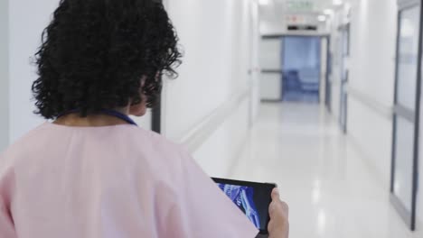 médico biracial enfocado caminando y usando una tableta en el hospital en cámara lenta