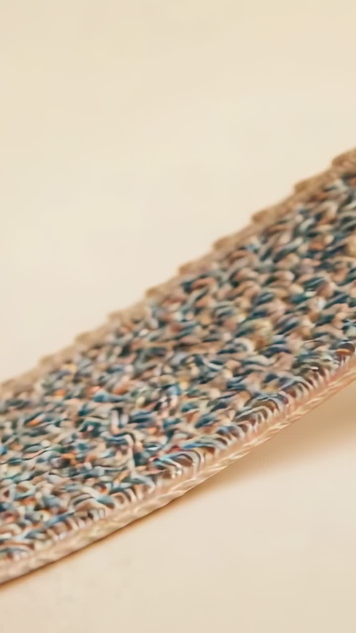 Video verticale: Un tappetino intrecciato che si appiattisce sul tavolo, rivelando filati colorati e un bordo intrecciato.