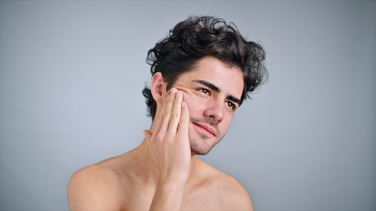 A young caucasian man with stubble beard is moisturizing face and skin with cosmetic oil while doing a facial massage. Slow motion