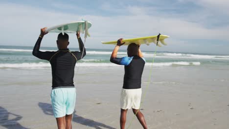 padre afroamericano y hijo adolescente de pie en la playa sosteniendo tablas de surf en la cabeza y hablando