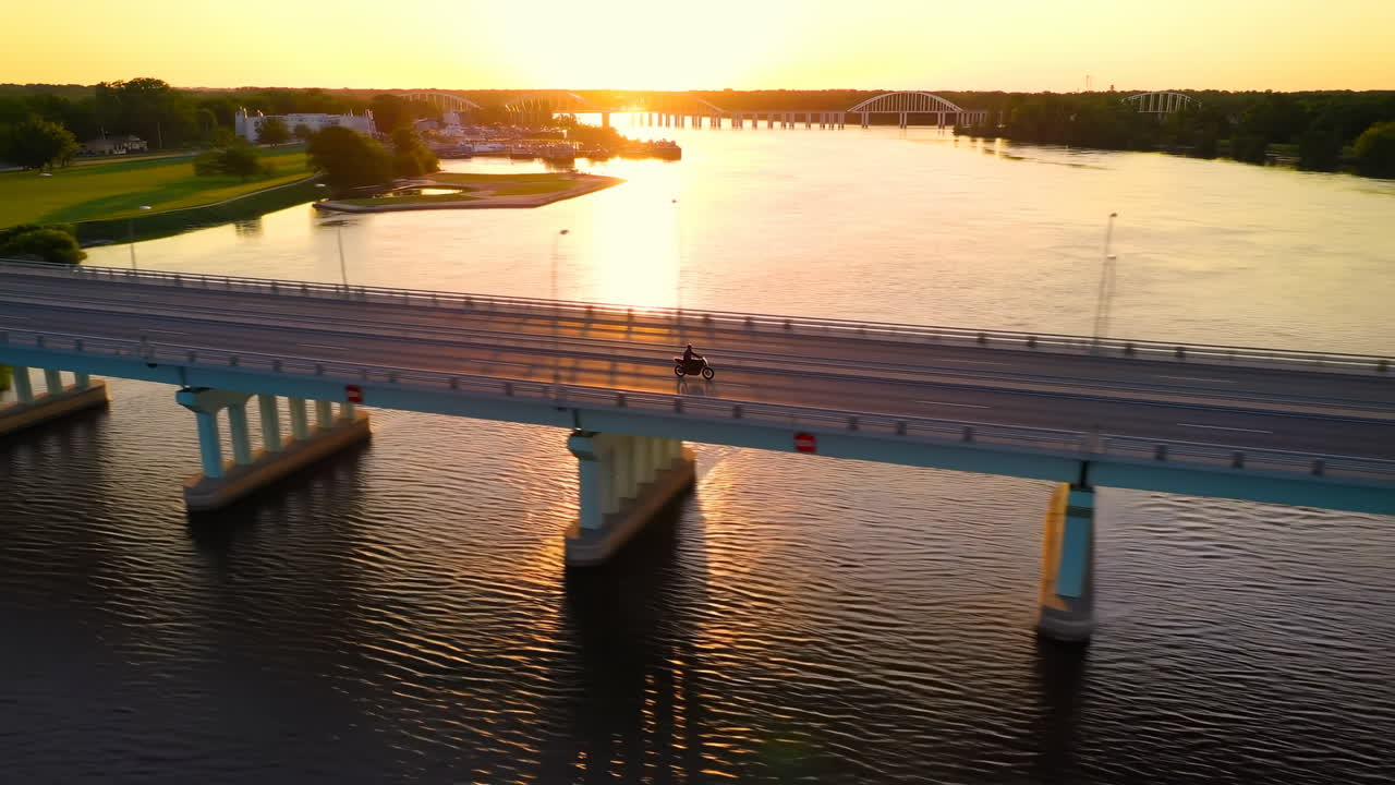 Ponte lungo al tramonto visto dall'alto con un ciclista che attraversa il fiume all'ora d'oro, luce solare calda che si riflette sull'acqua.