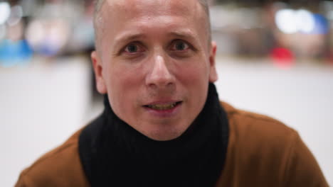 close-up of a middle-aged bald man laughing heartily, dressed in a brown sweater and black scarf, with a blurred background of people ice skating