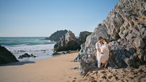 gentle pregnant couple posing on rocky beach leaning on rough stones hugging.