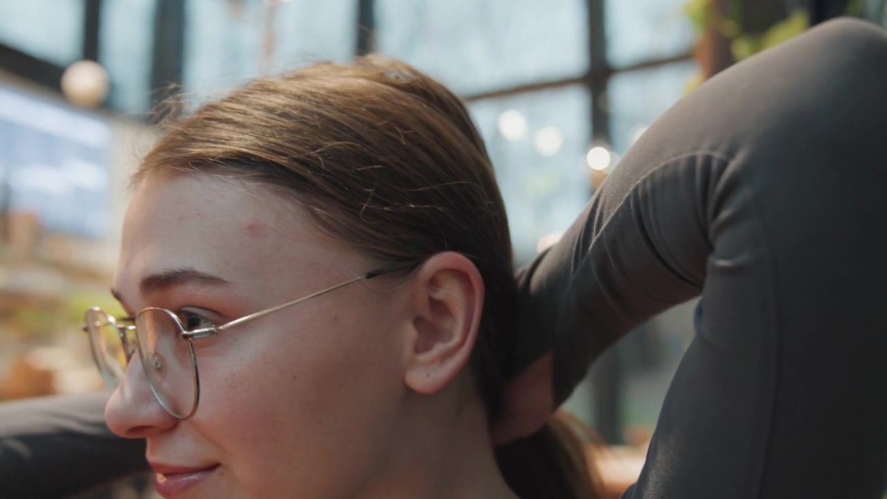 Giovane donna caucasica che si sistema la coda di cavallo in un affollato mercato coperto, con gli occhiali, le mani che le lisciando i capelli, piante e luci sullo sfondo, energia rilassata e sicura, una rapida routine per sistemarsi prima di un'apparizione pubblica.
