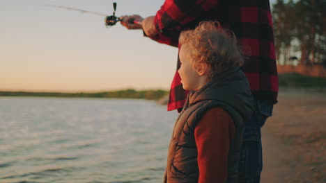 Neugieriges-Kleinkind-Beobachtet-Die-Natur,-Während-Sein-Vater-Bei-Sonnenuntergang-Eine-Glückliche-Familie-In-Der-Natur-Fischt