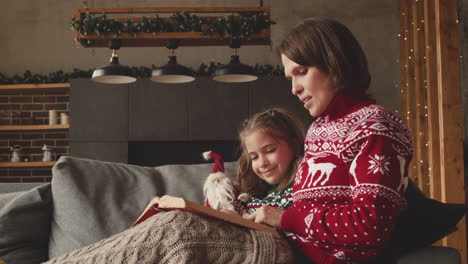 lovely mother sitting on couch with adorable daughter and reading her fairy tale on christmas day at home 3