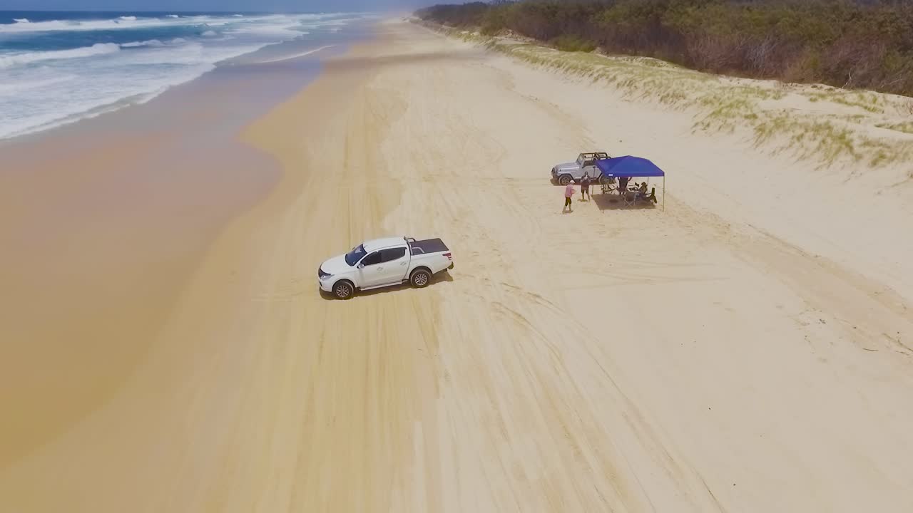 Low aerial drone shot travelling South down Main Beach on Queensland's North Stradbroke Island towards a camp site, with 4x4 four wheel drive vehicles driving on the beach