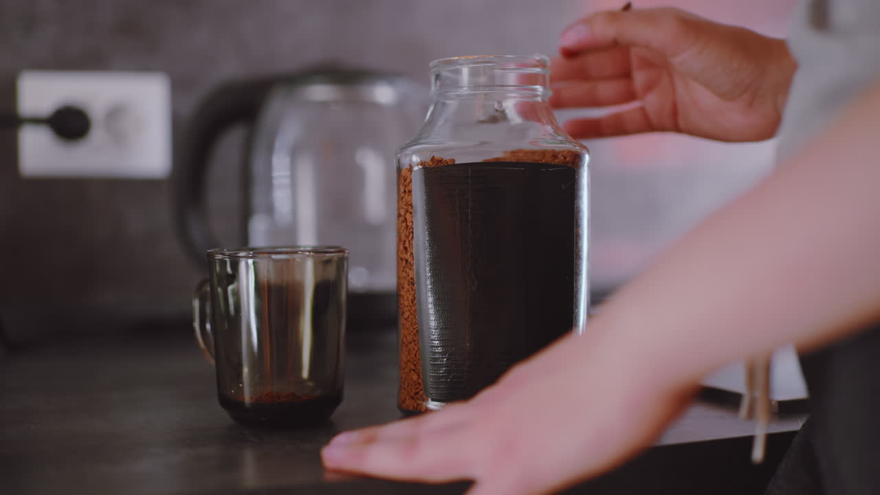 Close up of hand holding coffee container while water heats inside electric kettle, steam rising gently, person begins to turn content into cup, preparing warm drink during quiet kitchen moment