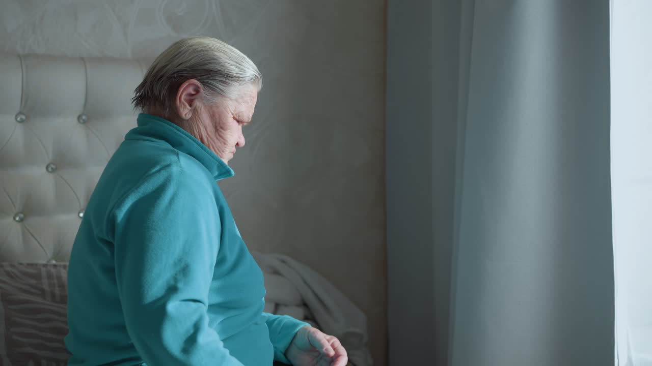 Elderly woman in green jacket bending forward with hand on lower back, appearing to ease discomfort, standing near window in bright, soft lighting, home setting with relaxed atmosphere