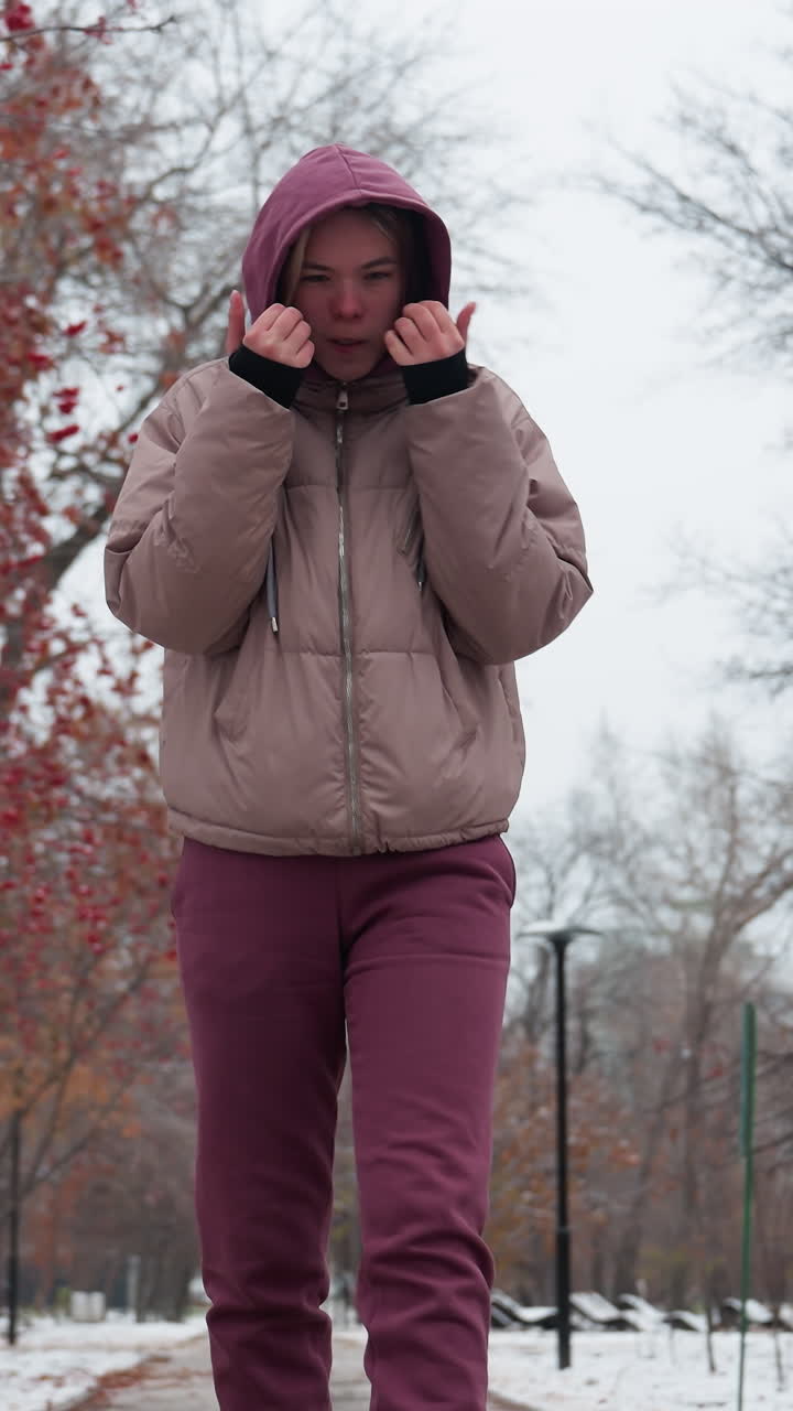 mujer joven vestida con ropa cálida de invierno camina a través de un parque nevado, sosteniendo sus manos cerca de su cara para el calor, árboles desnudos, bayas rojas, y figuras distantes
