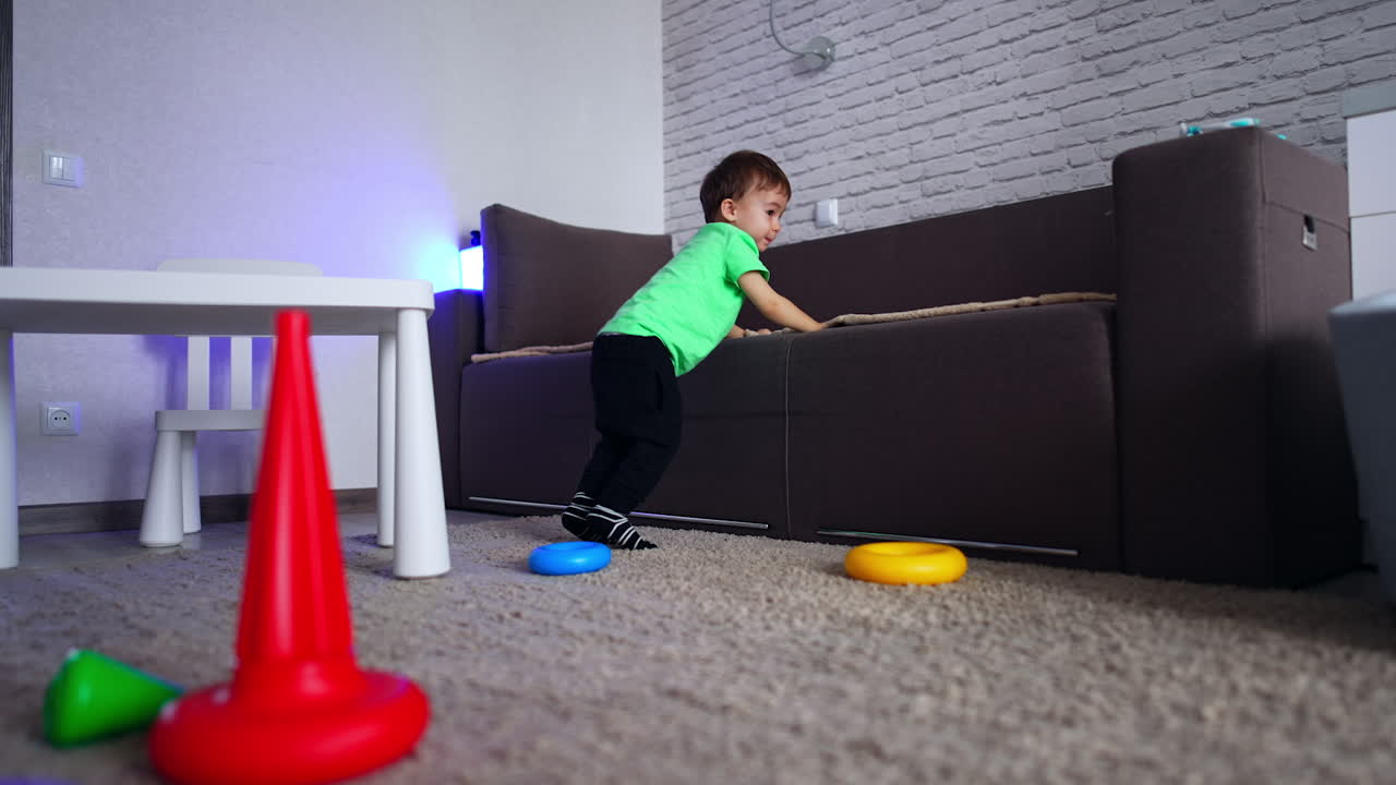 Energetic Caucasian toddler standing at the sofa jumping cheerfully. Baby boy pulling up his pants approaching camera. Low angle view.