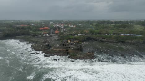 grey overcast aerial orbits tanah lot temple as waves crash on shore