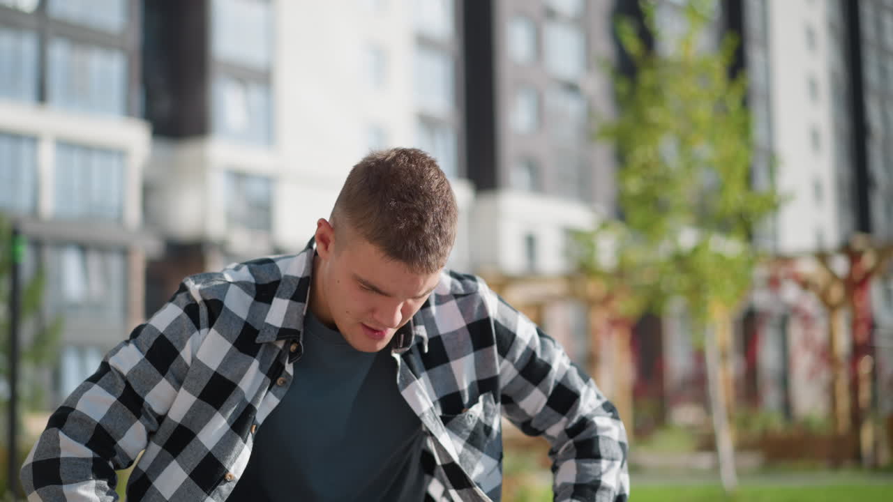 young man in checkered shirt bends forward while retrieving medicine from pocket with distant gaze, seated in outdoor park surrounded by greenery and tall residential buildings in blur background