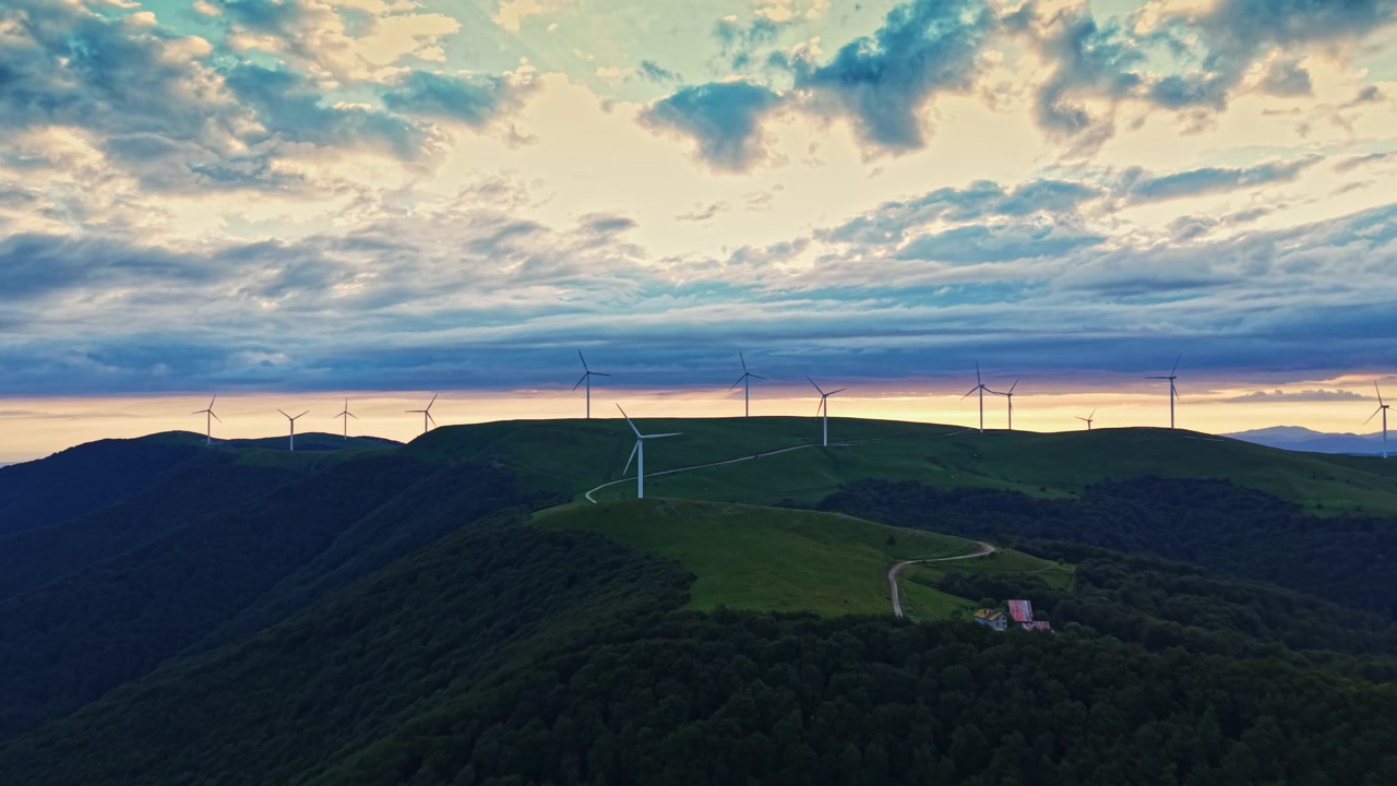 Turbine eoliche si ergono su una collina verde sotto un cielo nuvoloso al tramonto in una zona rurale.