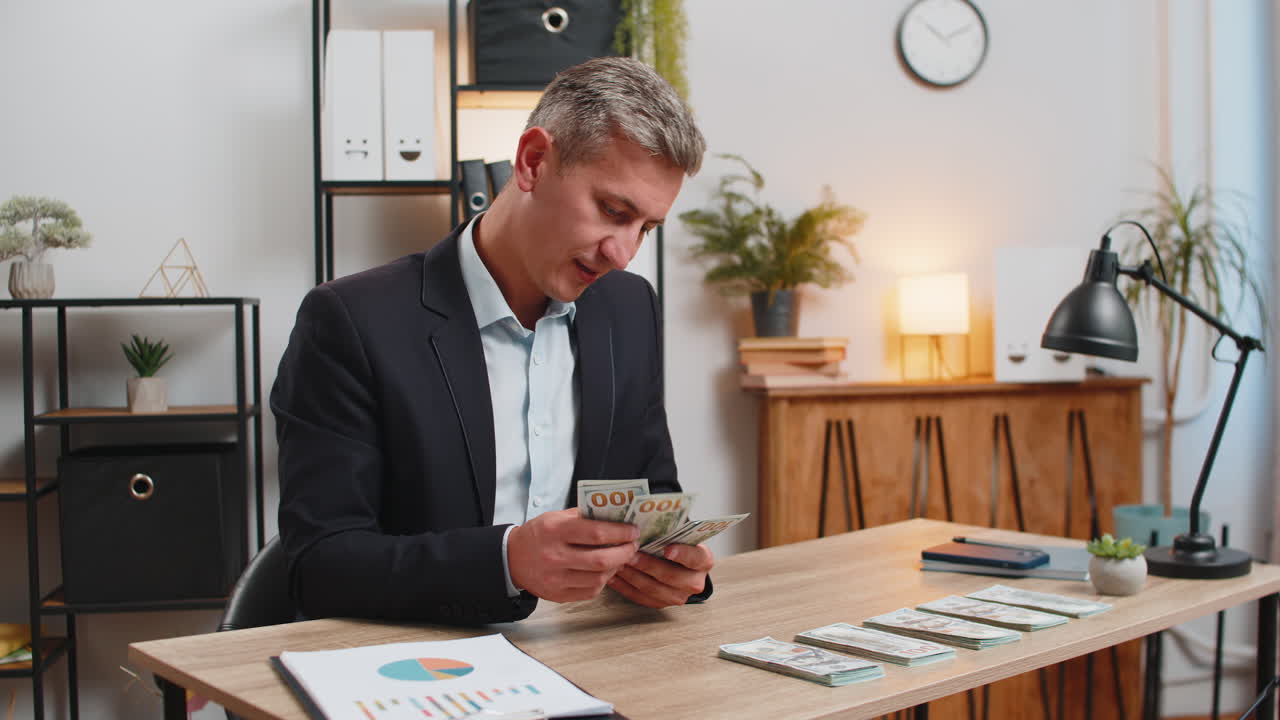 счастливый молодой белый успешный бизнесмен в формальном костюме подсчитывает деньги наличные в домашнем офисе стола