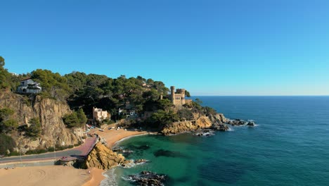 Aerial-views-of-the-main-beach-of-Lloret-De-Mar,-Castle-in-the-background,-wide-beach-without-people,-thick-sand,-water,-turquoise-blue