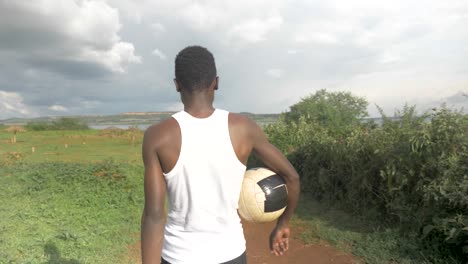 behind view tracking a young african man as he walks towards lake victoria with a football under his arm