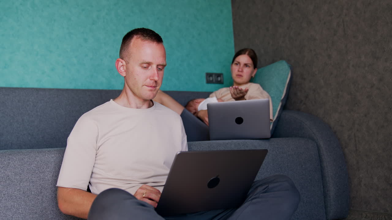 Woman with a child in hands sits on sofa. Wife talks to her husband about their work on laptop.