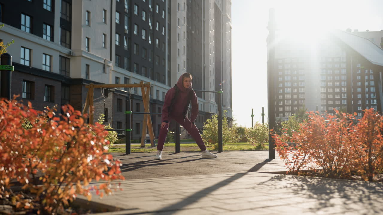 Long shot of wellness minded woman in hoodie and jacket performing side lunge exercise with hands on legs in modern urban park setting framed by flower beds under warm sunlight and autumn ambience