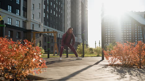 long shot of wellness minded woman in hoodie and jacket performing side lunge exercise with hands on legs in modern urban park setting framed by flower beds under warm sunlight and autumn ambience