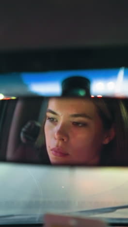 a woman is seen in the rearview mirror of her car as she waits in traffic, with the reflection of another car visible behind her through the rearview mirror and blurred city lights in the background