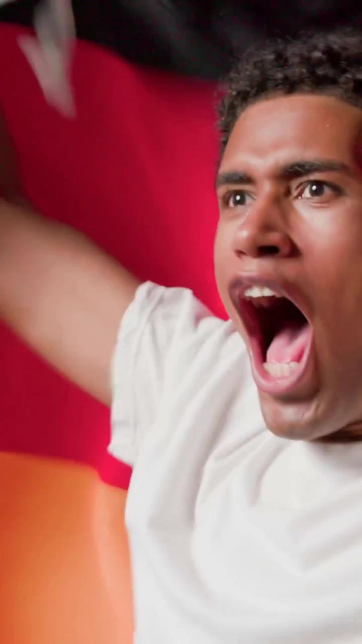 Vertical video: Smiling man cheering, pulling jacket showing white T studio, waving red-orange flag