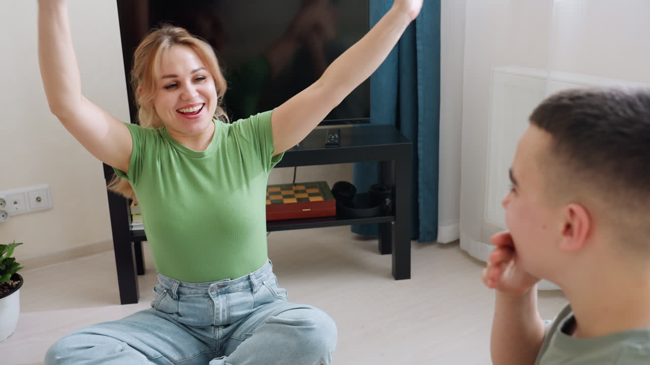 Smiling mother enjoys joyful moment playing hand game with son while sitting barefoot on clean wooden floor in bright living room with indoor plant and TV on black stand