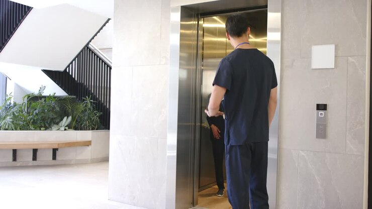 Diverse med staff calling elevator at lobby, woman emerging, man showing tablet, discussing care