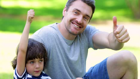 padre sentado con el hijo pequeño en el parque mostrando los pulgares hacia arriba