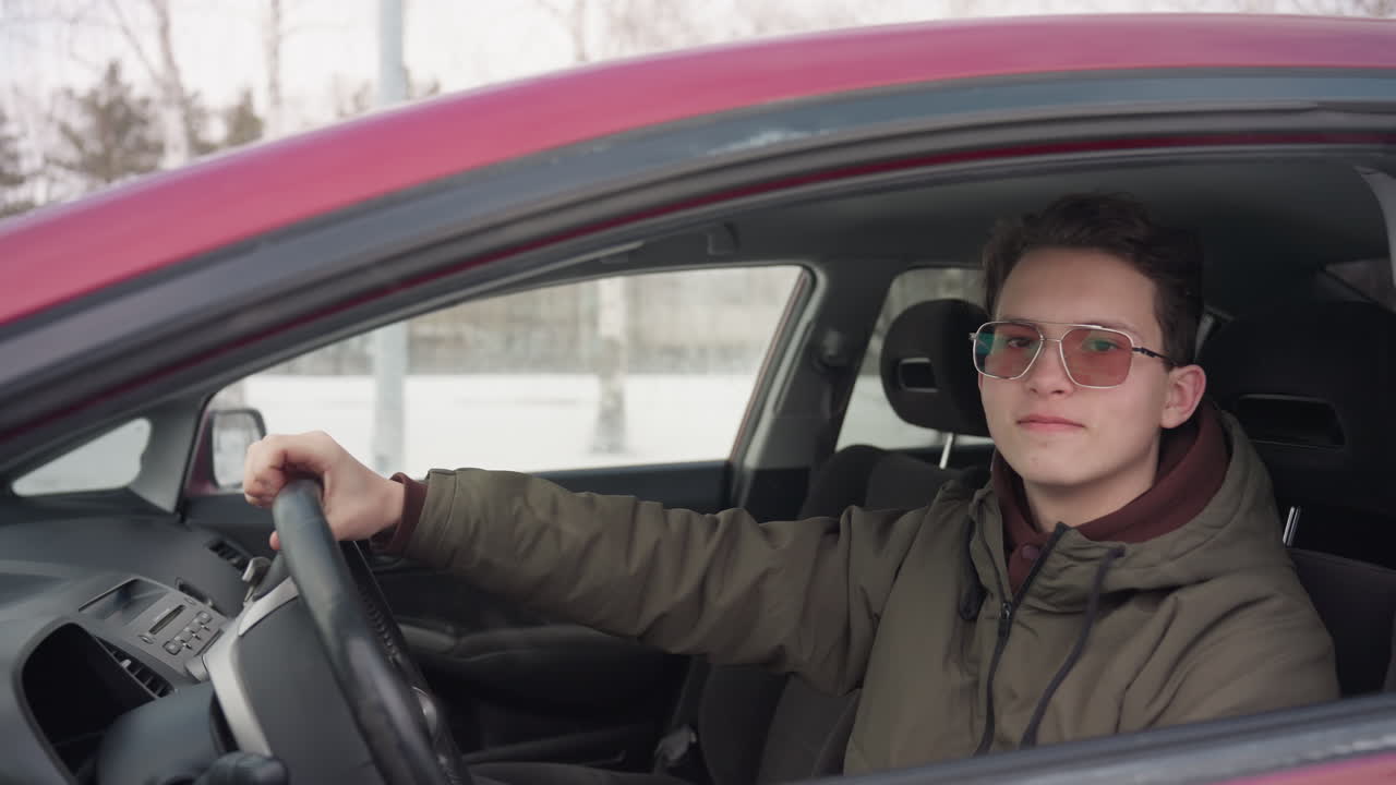 white teenage boy wearing winter jacket sitting in parked red car holding steering wheel with one hand smiling confidently while distant car passes by blurred in snowy outdoor background