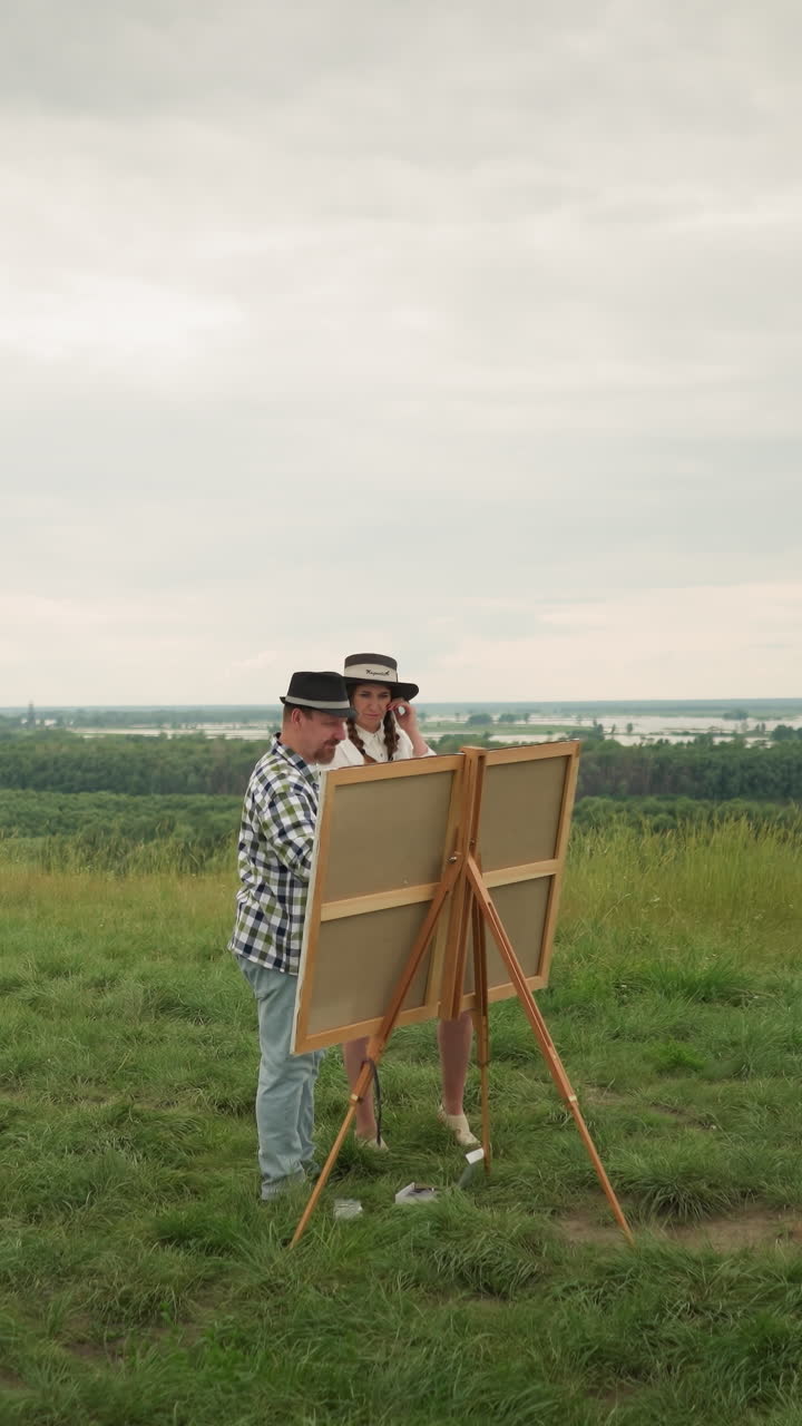 A man in a black hat and plaid shirt stands close to a woman in a white dress, engaged in a discussion or observation in a grassy field with a drawing board set up before them. with a river