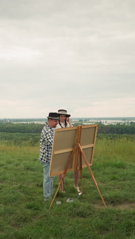 a man in a black hat and plaid shirt stands close to a woman in a white dress, engaged in a discussion or observation in a grassy field with a drawing board set up before them. with a river