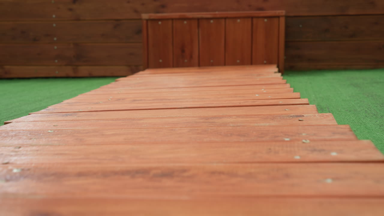 Trægangbro, der fører til dekoreret baldakin. Grønt græstørv flankerer patinerede planker, rent rødt draperi og hjerteornamenter med løv skaber en intim kulisse til valentinsdag eller bryllup, lav vinkel.