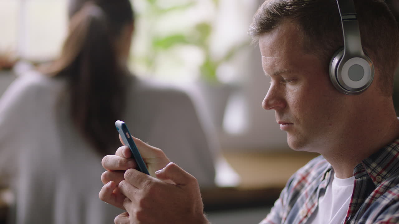 카페에서 스마트폰을 사용하는 젊은 백인 남자가 문자 메시지를 입력하고 소셜 미디어에서 아이디어를 공유하고 헤드폰을 착용하여 음악을 듣는 것을 즐기고 있습니다.