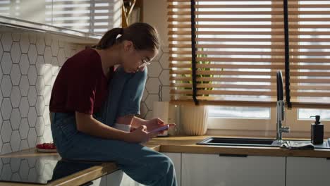 brede opname van een blanke tienermeisje die in de keuken zit en in stilte een mobiele telefoon gebruikt