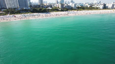 Aus-Der-Höhe-Eröffnet-Sich-Ein-Spannender-Panoramablick-Auf-Die-Türkisfarbene-Wasseroberfläche,-Hinter-Der-Sich-Ein-überfüllter-Strand-Erstreckt,-Und-Dahinter-Sind-Große-Metropolen-Mit-Hochhäusern-Zu-Erkennen