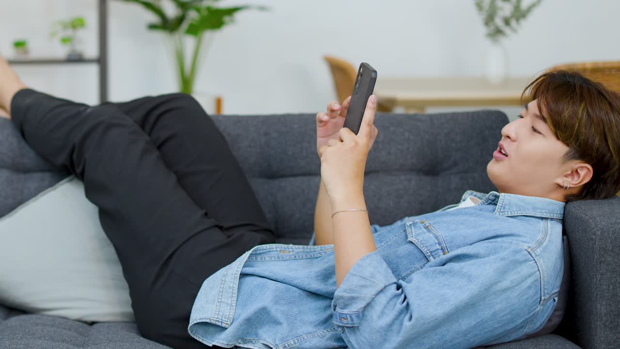 Un giovane asiatico è sdraiato su un divano grigio e guarda il suo smartphone a casa.