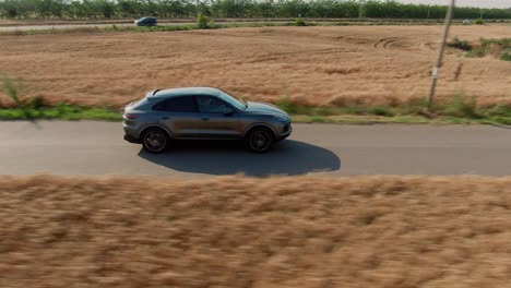 aerial side shot of a grey luxury suv sport car accelerating on a country tarmac road surrounded by golden wheat fields