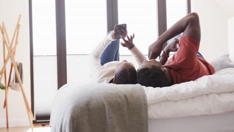 happy african american couple lying on bed using smartphone and talking at home, in slow motion