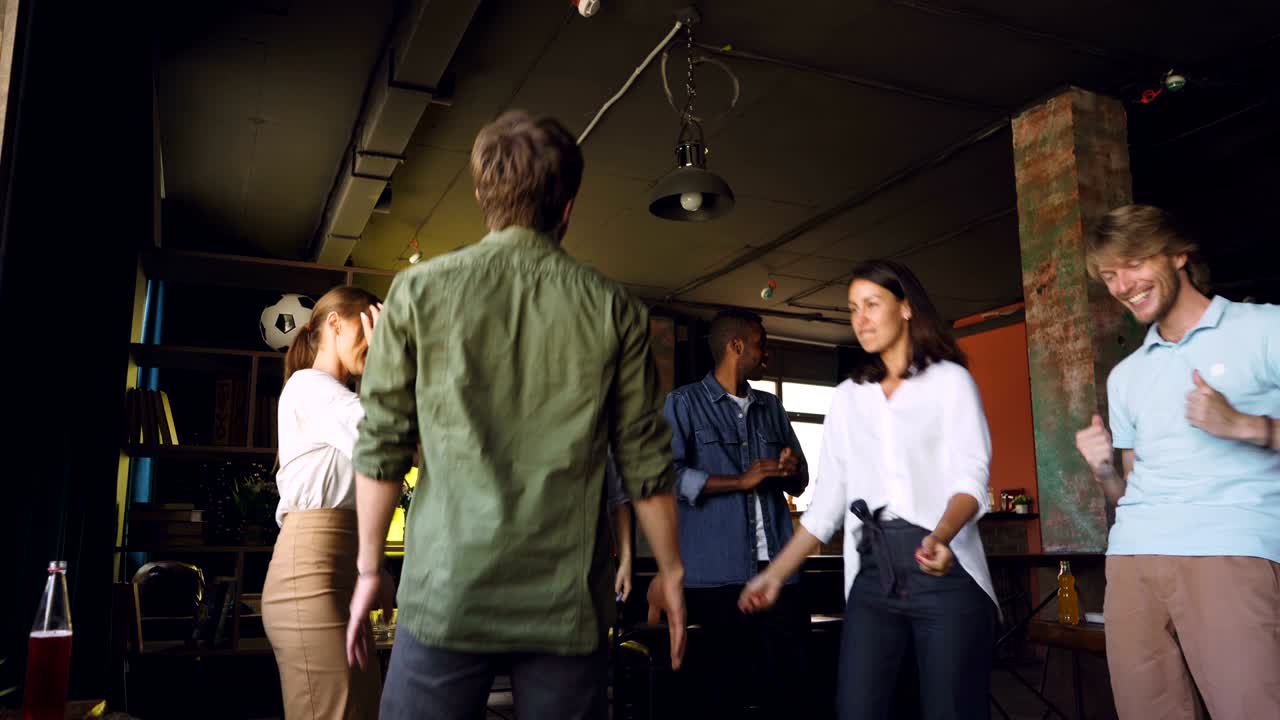 Happy young people are dancing in circle at office party, clapping hands and laughing enjoying corporate holiday. Men and women are wearing casual clothing.
