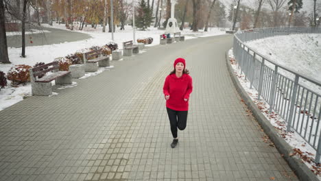 mujer corriendo por el camino nevado del parque con capucha roja y leggings negros, rodeada de un sereno paisaje de invierno con bancos, arbustos, barandillas de hierro, postes de lámparas y un distante monumento a la cruz