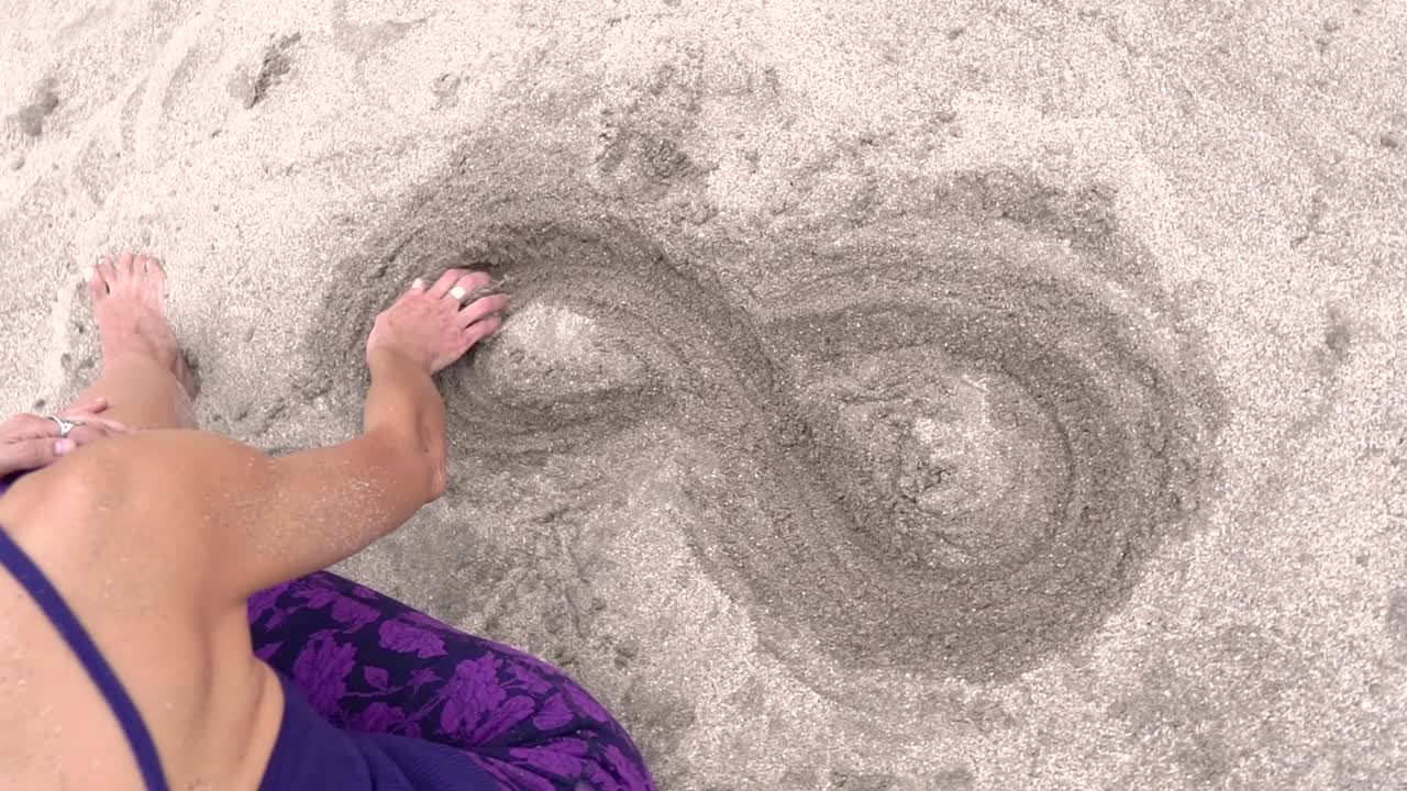 A high-angle view of a woman on a beach, she is doing the infinity symbol on the sand