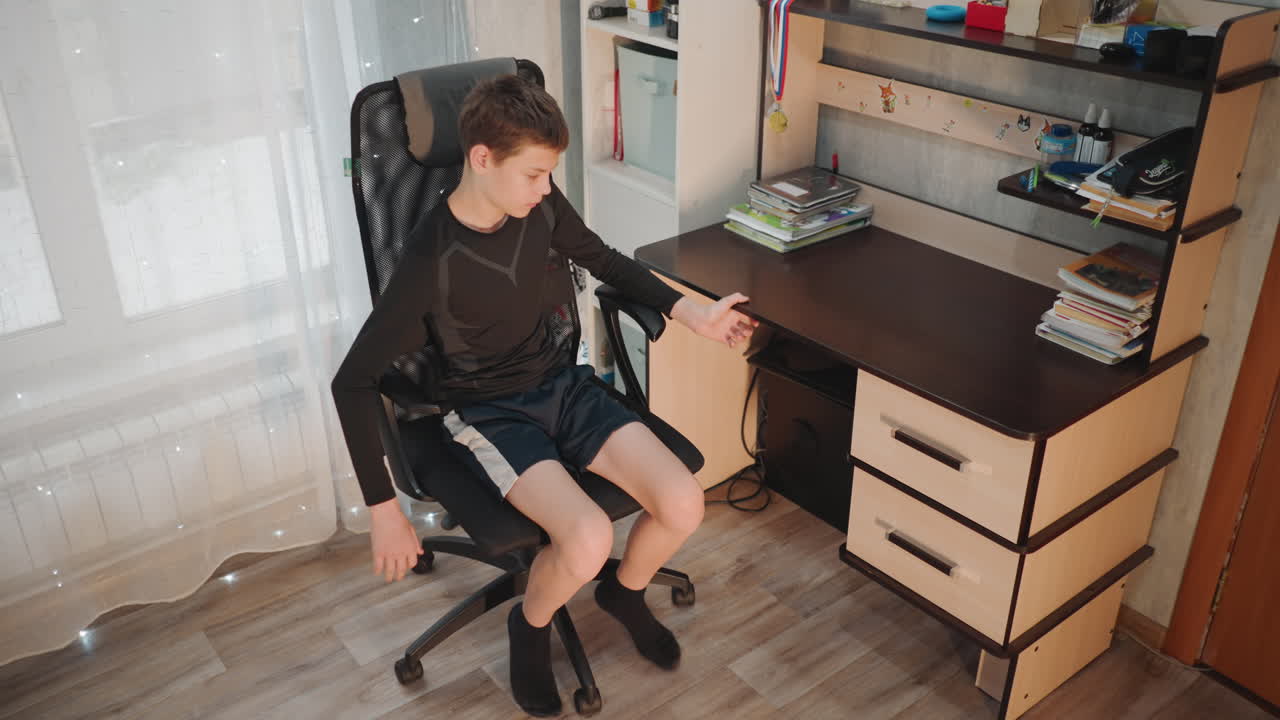 Juvenile male shifts while seated at desk. Adolescent male relaxes and moves in his study environment. Teenager adjusts and shifts position in swivel chair at his personal study space
