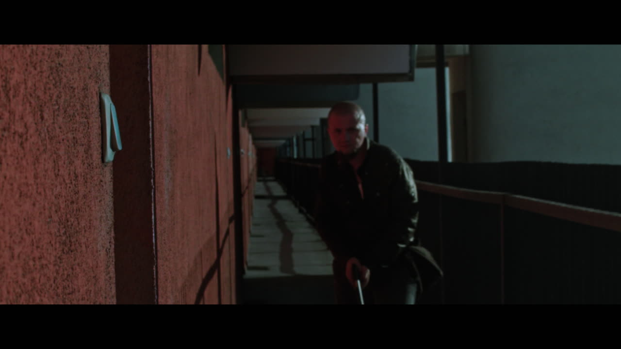 A bald dangerous looking man enters the outside corridor and shoots a gun