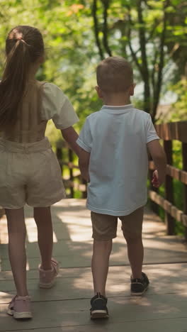 little girl and boy hold hands walking together along viaduct in forest. happy children enjoy travel to shady summer park. kids explore nature jumping