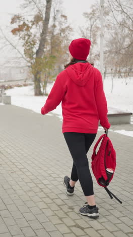 athlete wearing red jacket with headphones around neck walking along interlocked pathway holding bag in left hand near iron railing in serene park with snowy ground and foggy atmosphere