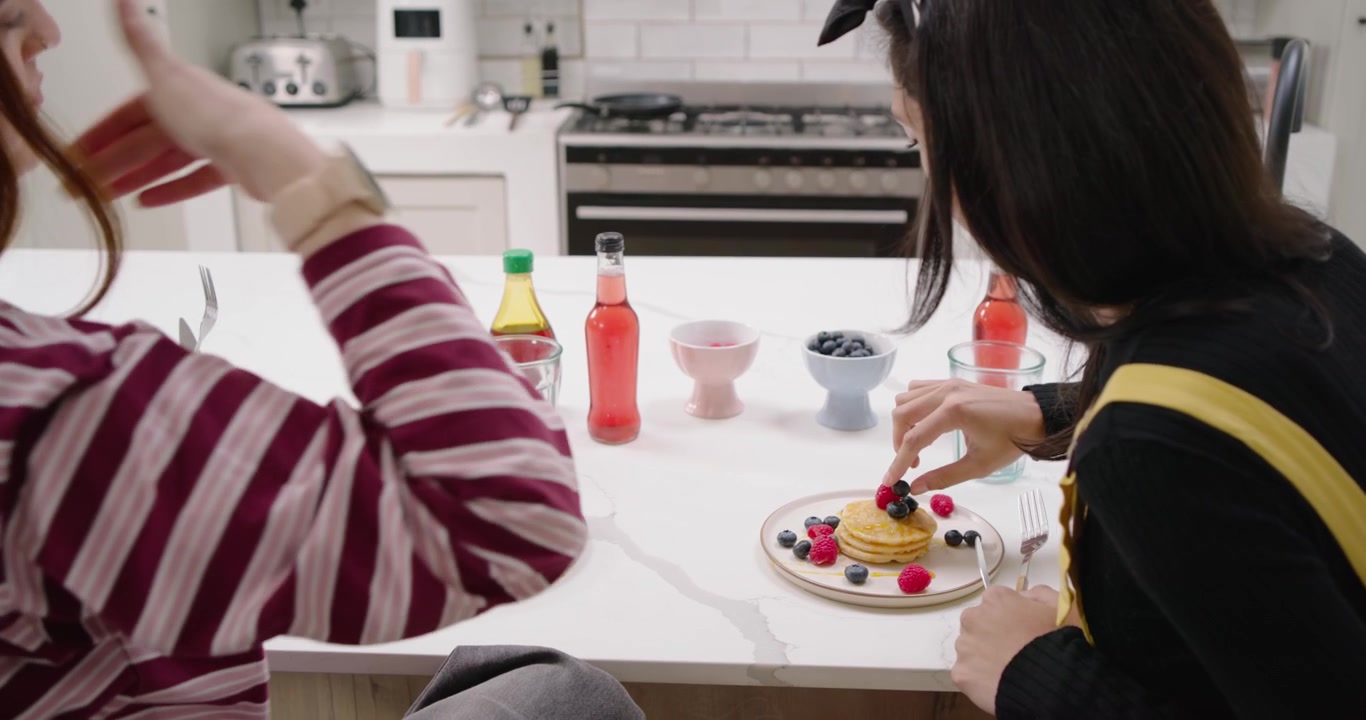 Due amiche sedute in cucina, la donna a destra prende i frutti di bosco, li aggiunge al pancake e prende le forchette.